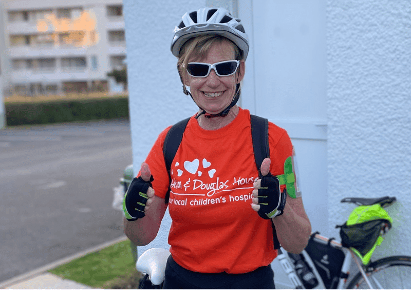 Janet smiling and giving two thumbs up. She is wearing a cycle helmet, sun glasses and an orange t-shirt with the Helen & Douglas House logo on it.