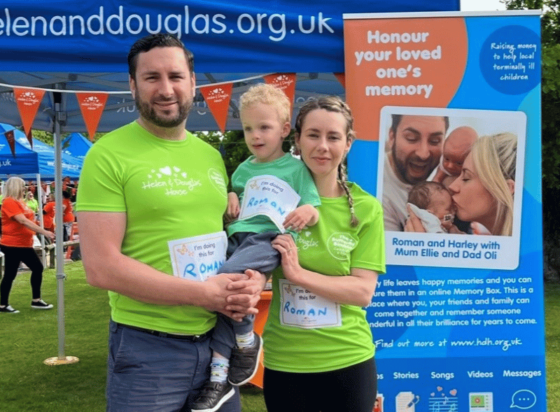 Eli, Oli and Harley at Butterfly Bimble Oli and Ellie holding their young son, Harley. They are standing in front of a Helen & Douglas House banner.