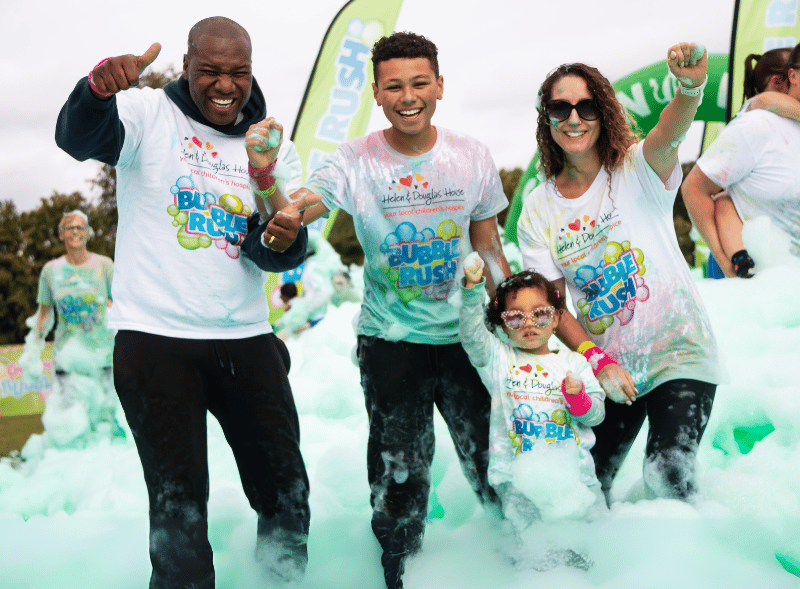 A mum, dad, teenage boy and young girl holding hands and cheering. They are standing in knee high green bubbles. 