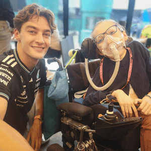 George Russell smiling and taking a selfie with a young girl in a power chair. She is wearing breathing apparatus. 