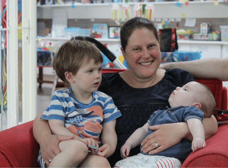 A mum smiling whilst holding her young son and a baby