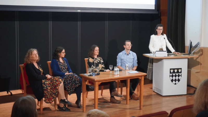 A panel of 3 women and a mad sat at a table on stage while a woman presents at a podium