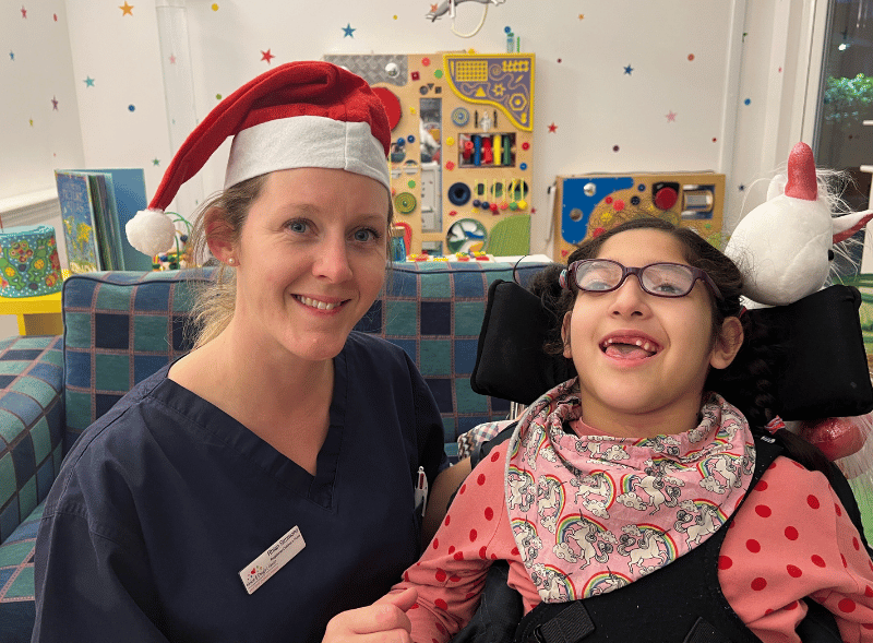 A female nurse smiling and wearing a Santa hat. She is next to a young girl in a wheelchair.